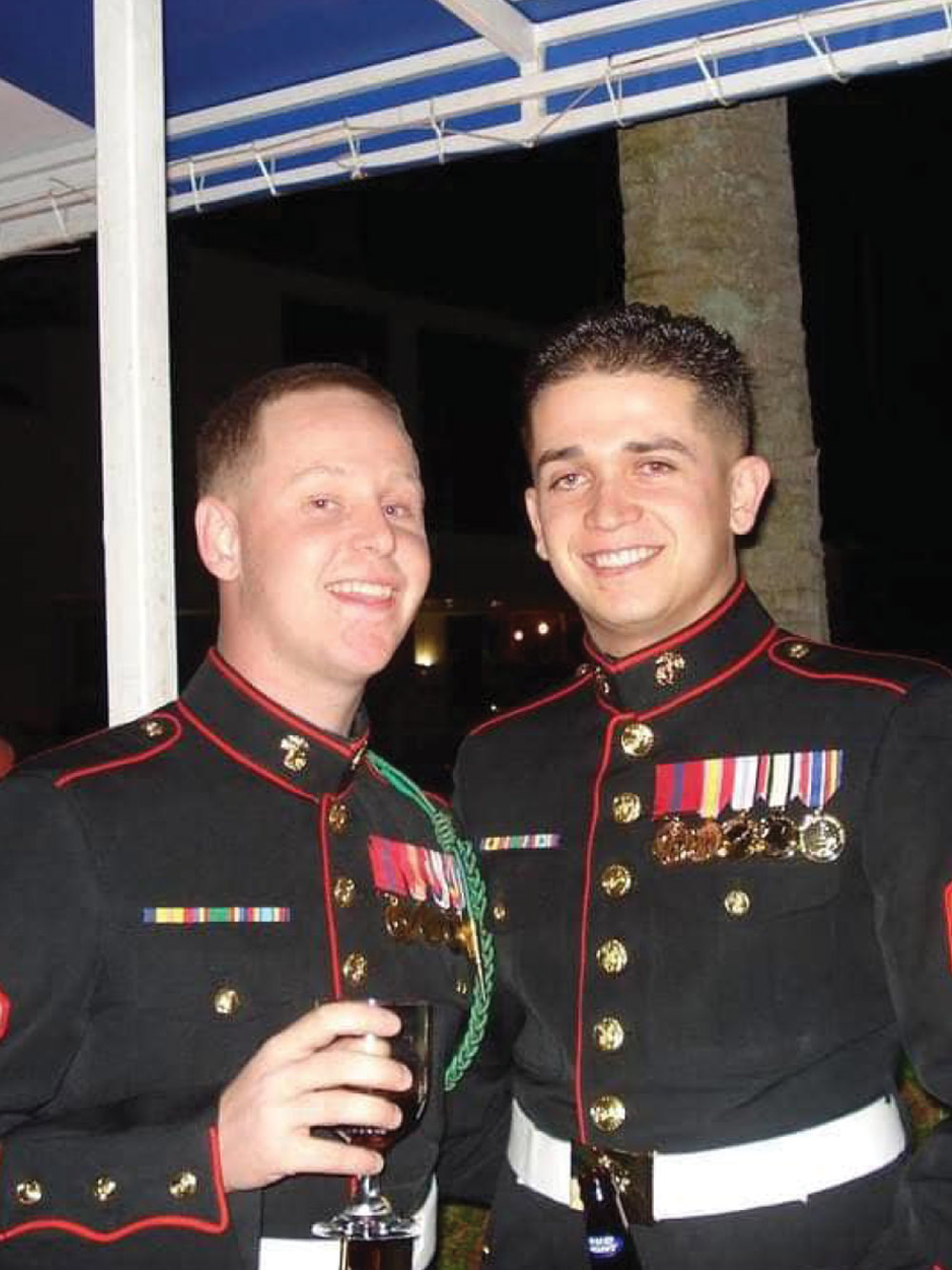 Two U.S. Marines are smiling and posing for a photo at a formal event. They are both wearing their dark blue dress uniforms with red trim, medals, and a white belt. The Marine on the left is holding a glass of wine.