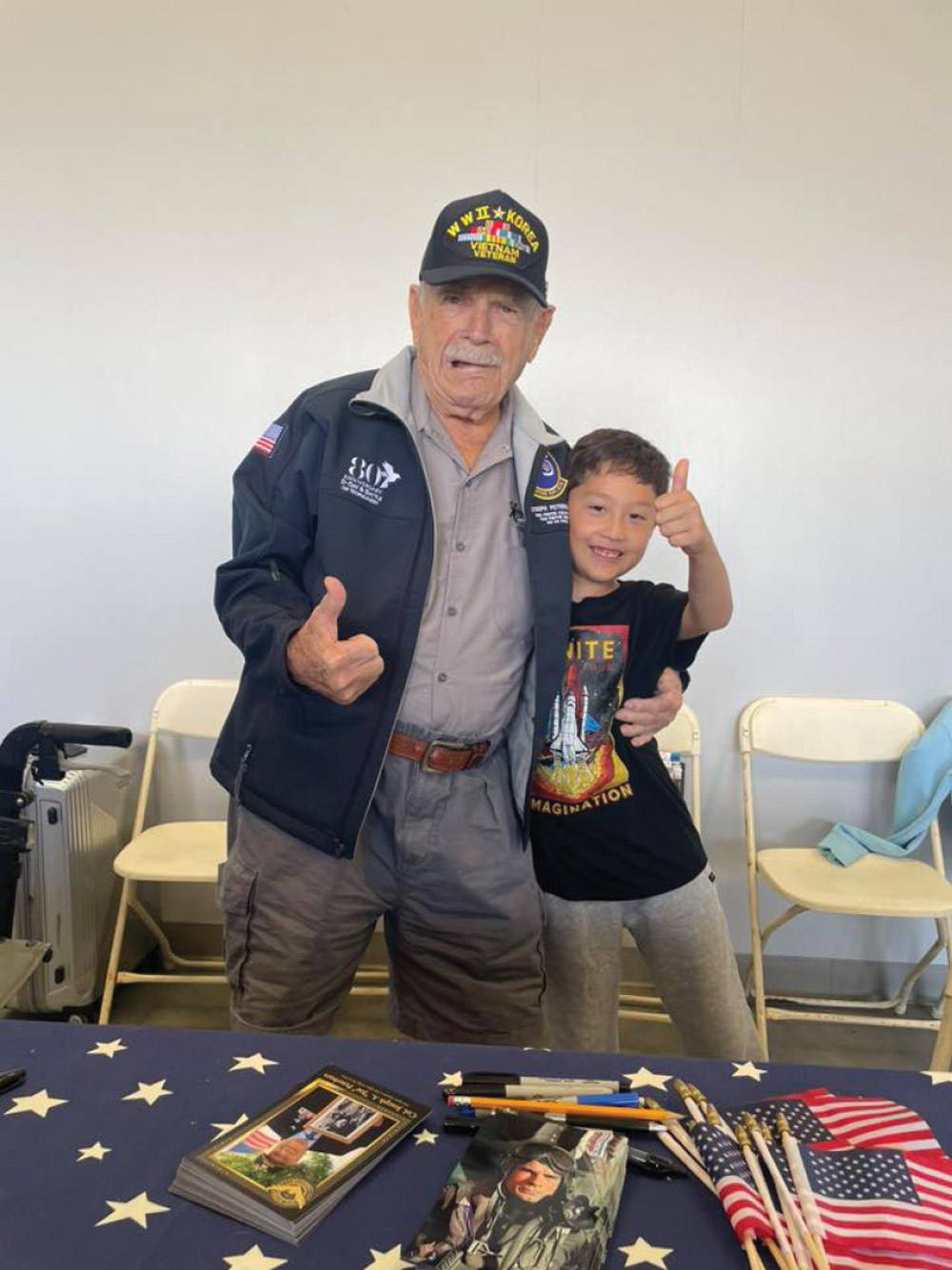 A photo of a veteran and a young boy standing side-by-side. The veteran, an older man with a mustache and a hat that reads "W.W. II + KOREAN + VIETNAM VETERAN," is giving a thumbs-up to the camera. The young boy, standing next to him with his arm around the veteran, is also giving a thumbs-up. A table in front of them is covered with a star-patterned cloth and has photos and small American flags on it.