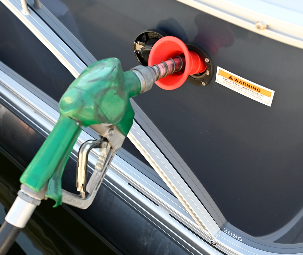 An overhead close-up shows a green fuel nozzle inserted into the fuel fill port of a boat, which has a dark gray hull. A bright orange funnel-like adapter is being used in the fuel opening. A "WARNING: FUEL SYSTEM UNDER PRESSURE" sticker is visible on the hull nearby.