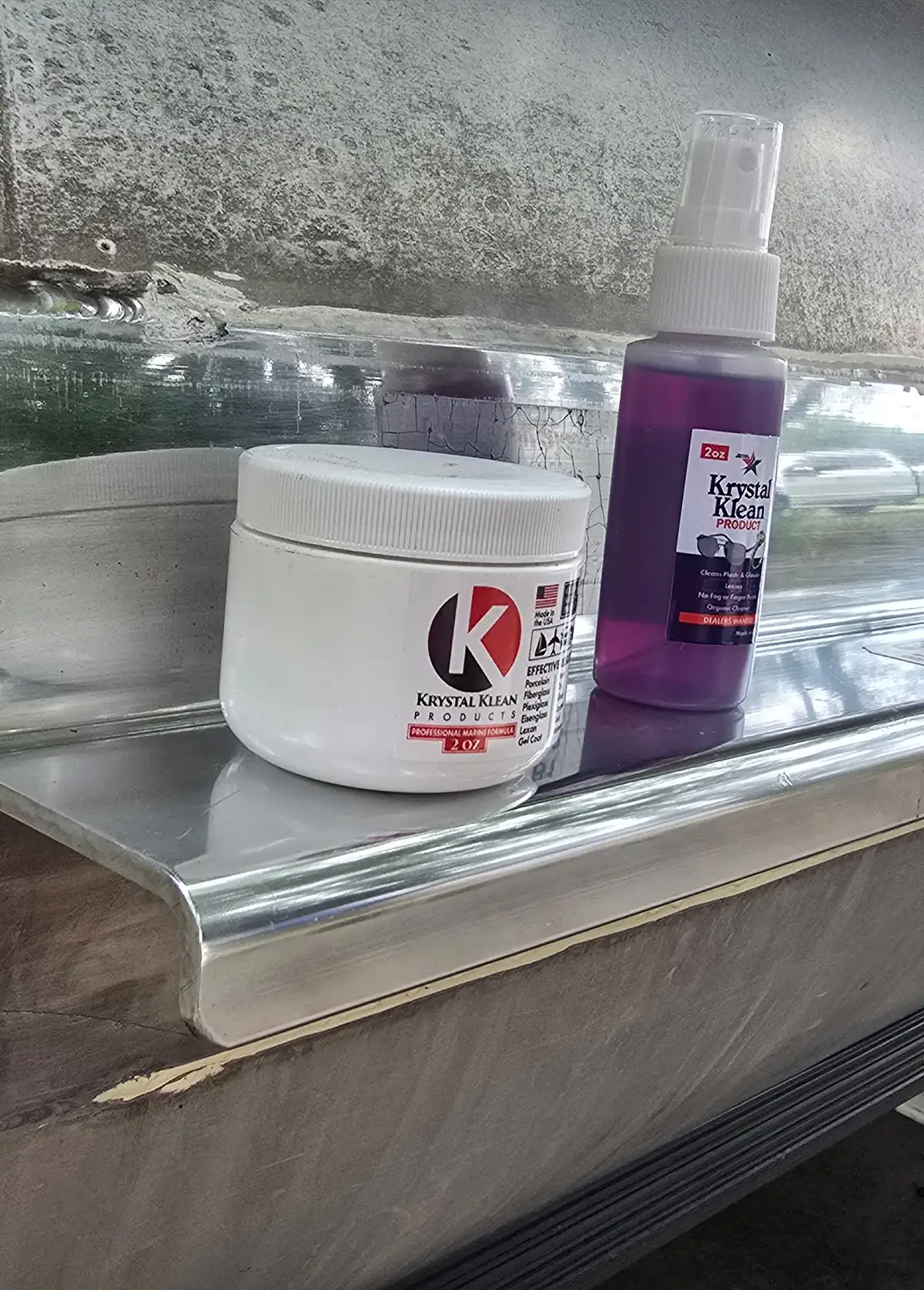 Two cleaning products from Krystal Klean Products are displayed on a metallic surface, likely a boat trim. One is a white jar labeled with 'Professional Grade Polish/Sealant 4 OZ,' and the other is a purple spray bottle with 'Krystal Klean Products, 2 OZ' for cleaning or protection.