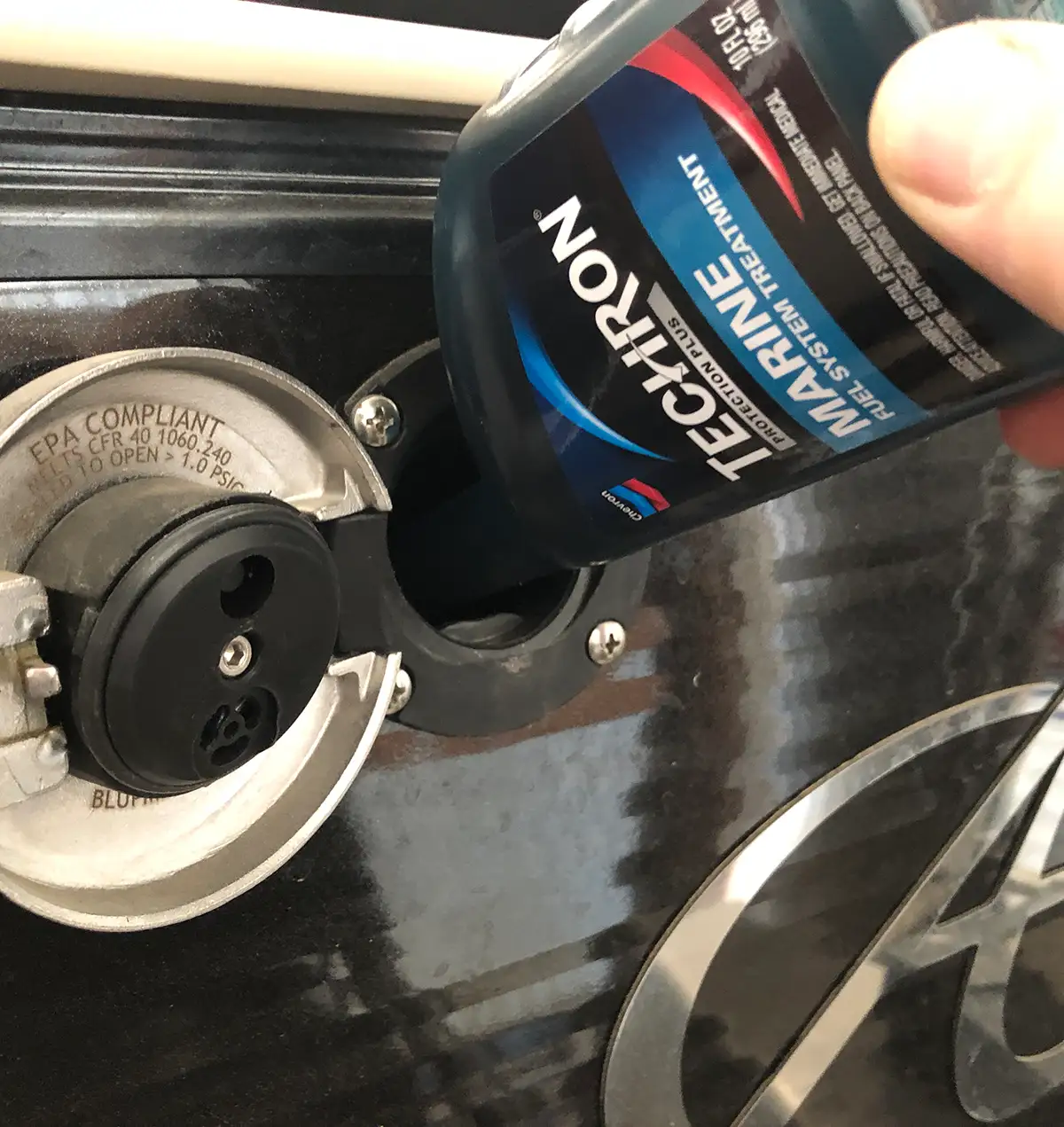 An overhead close-up shows a hand pouring Techron Marine Fuel System Treatment from a dark bottle into the fuel filler neck of a boat. The boat's fuel cap is open and labeled "EPA COMPLIANT". The surrounding surface of the boat is black and glossy.