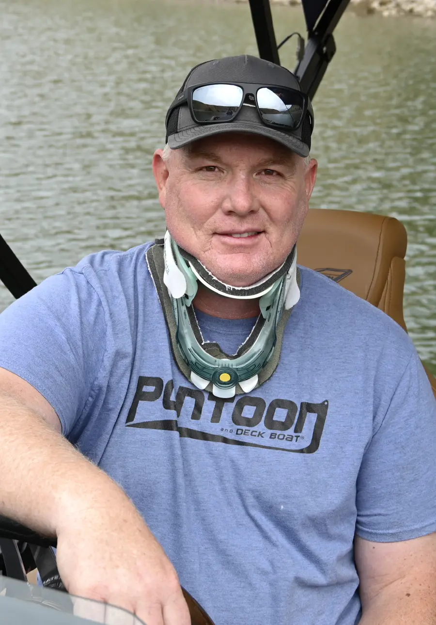 Portrait orientation close-up outdoor photograph of Brady L. Kay, a smiling man wearing a dark gray colored hat with black colored sunglasses on top and a neck brace wrapped around his neck as he is seated in the captain's chair of a pontoon motorboat looking toward at the camera; He is wearing a faded dark blue colored graphic t-shirt with the logo that reads PONTOON and DECK BOAT printed on it; The background is a lake with calm, murky water and a rocky shoreline