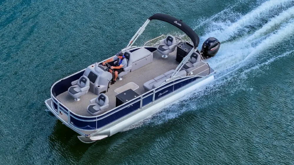 A landscape orientation cropped aerial photograph overhead view of a blue and silver colored Barletta Aria pontoon motorboat with a dark black canopy, moving quickly across a body of water and leaving a white wake behind it; A man is seated at the helm driving the vehicle; The pontoon motorboat has light gray seating with a contrasting darker gray trim