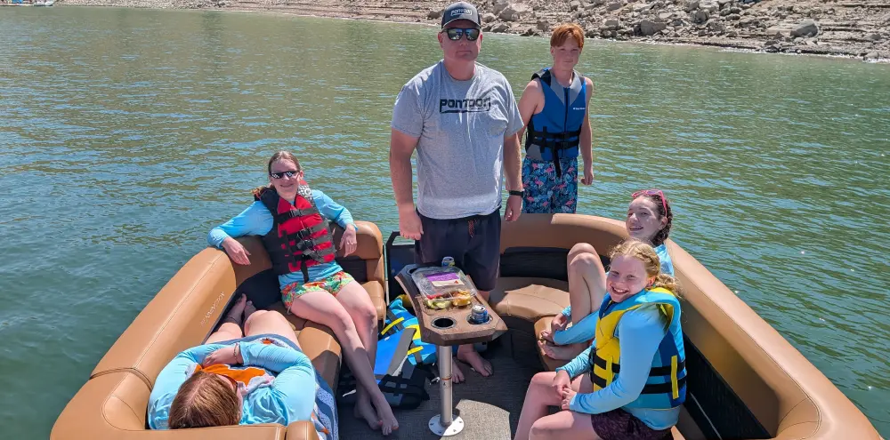 Landscape orientation cropped close-up outdoor photograph view of Brady L. Kay, a grinning man and his five children are posing for a photo on a pontoon motorboat on a sunny lake; Brady L. Kay is standing in the center, wearing black colored sunglasses, a dark grey colored Pontoon and Deck Boat graphic t-shirt, black swimming shorts, and a dark grey colored Pontoon and Deck Boat graphic logo hat; His children are seated around him on the brown leather seats of the pontoon motorboat, most wearing swimming life jackets and a rocky shoreline is visible in the background behind all of them