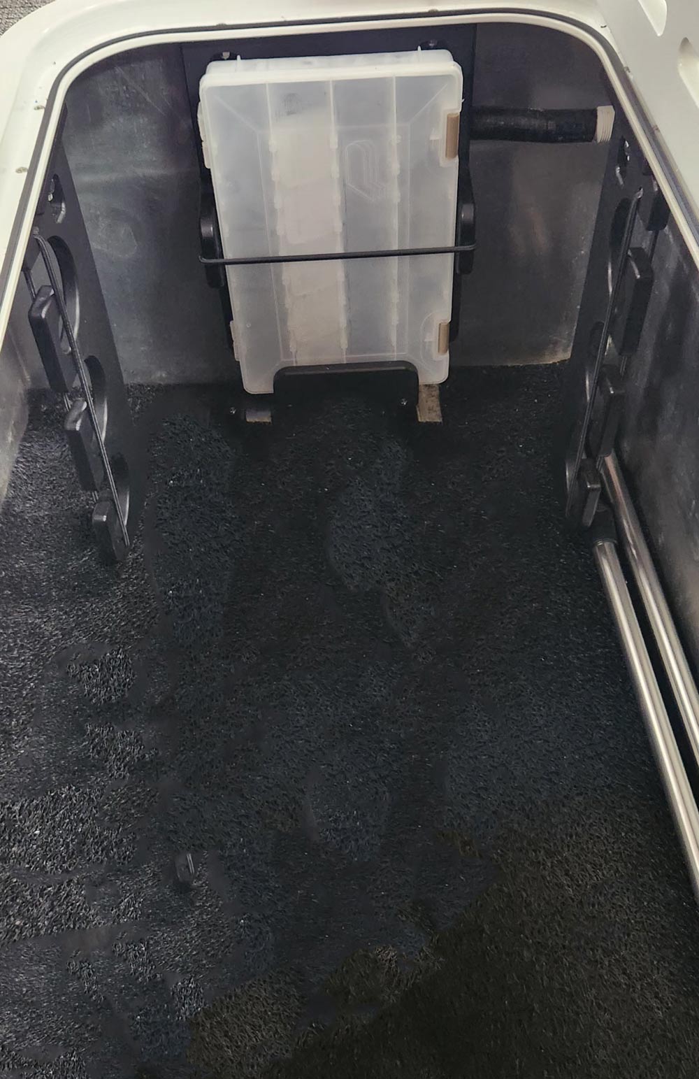 An open boat storage compartment with a black textured floor mat and a rectangular plastic tackle box secured inside.