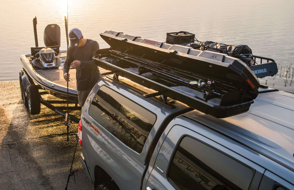 A fisherman standing on a dock next to his boat trailer, loading gear from a Yakima roof-mounted fishing rod box on the back of a pickup truck.