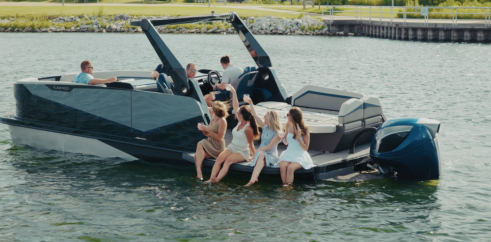 A modern boat with six people relaxing on calm water near a rocky shoreline.