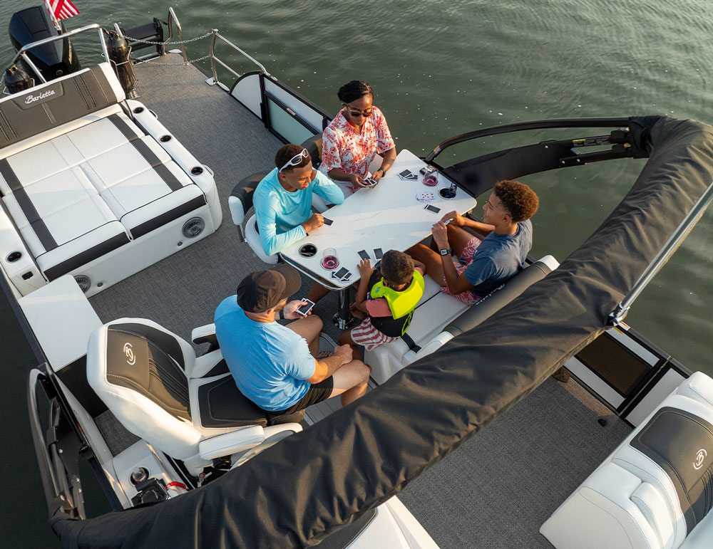 close aerial view of a family of five sitting around a Barletta pontoon passenger table and playing a card game as they float on water