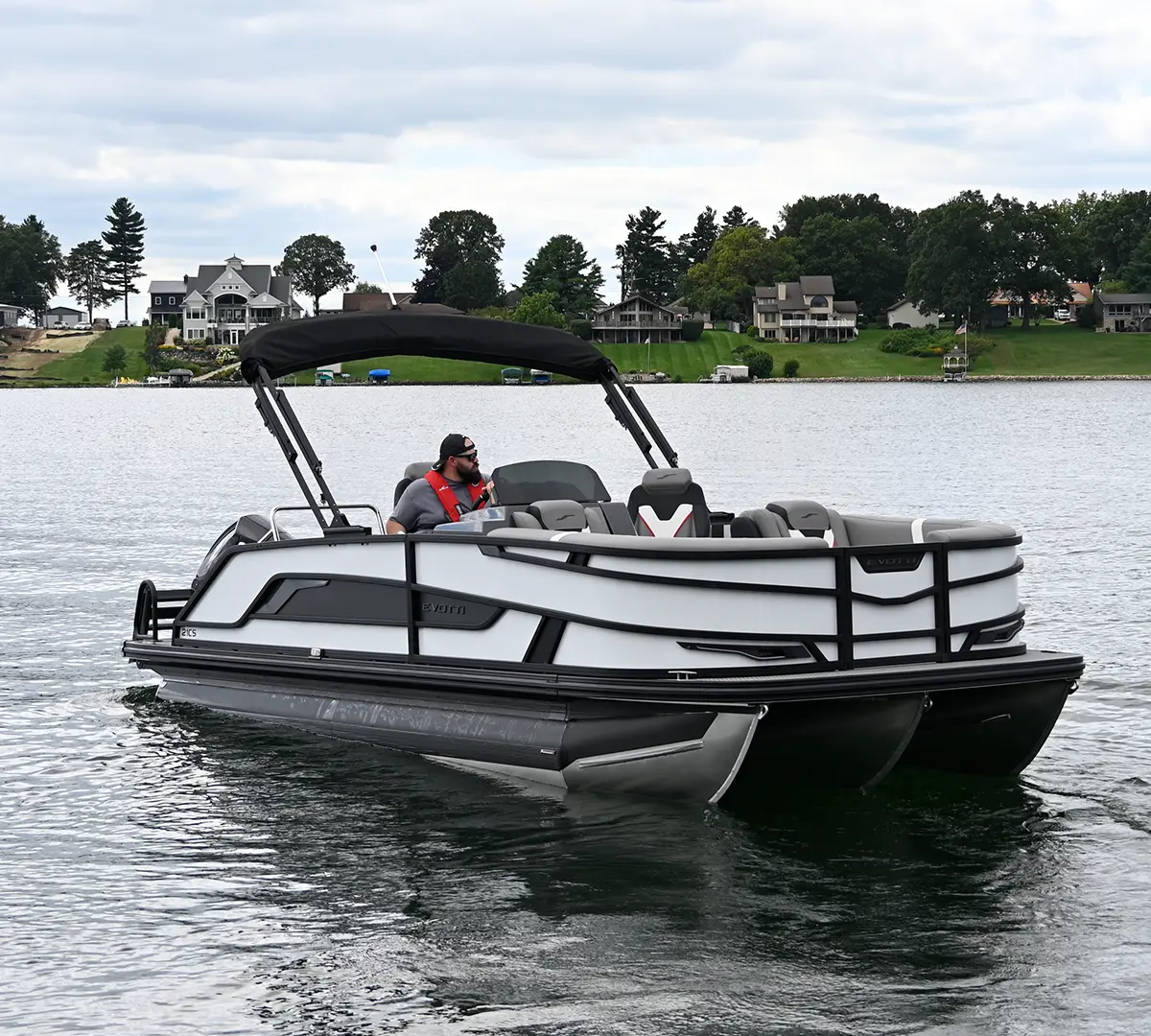 full view of a man riding at the helm of a black and white Evotti pontoon, a lakeside residential area lines the horizon in the background