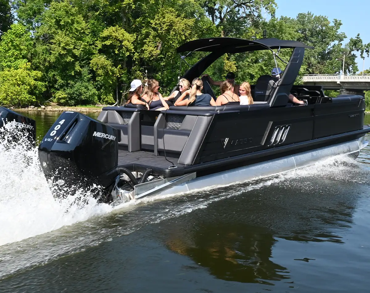 level view of a large group of women relaxing and mingling on an all black Viaggio pontoon sailing on a body of water lined by lush green trees