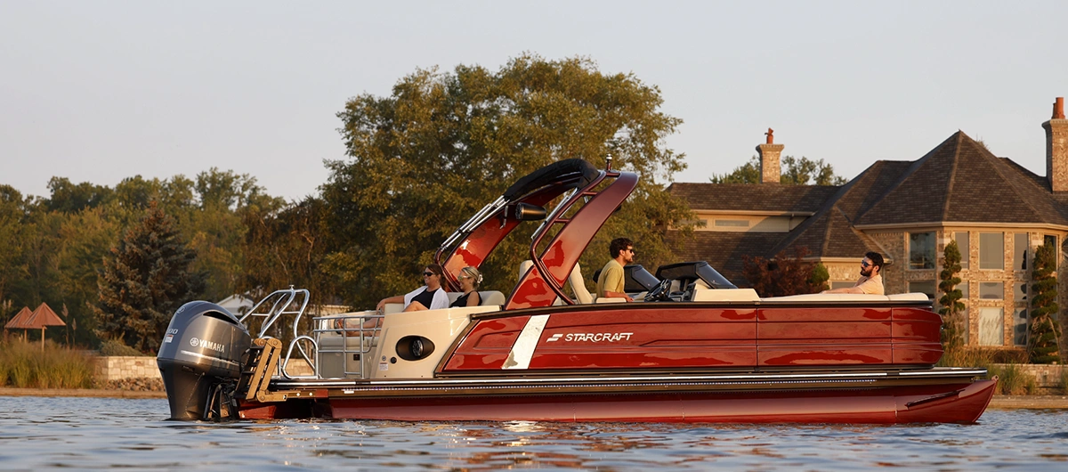 level starboard side view of a maroon Starcraft pontoon floating on a body of water beside a large residential structure