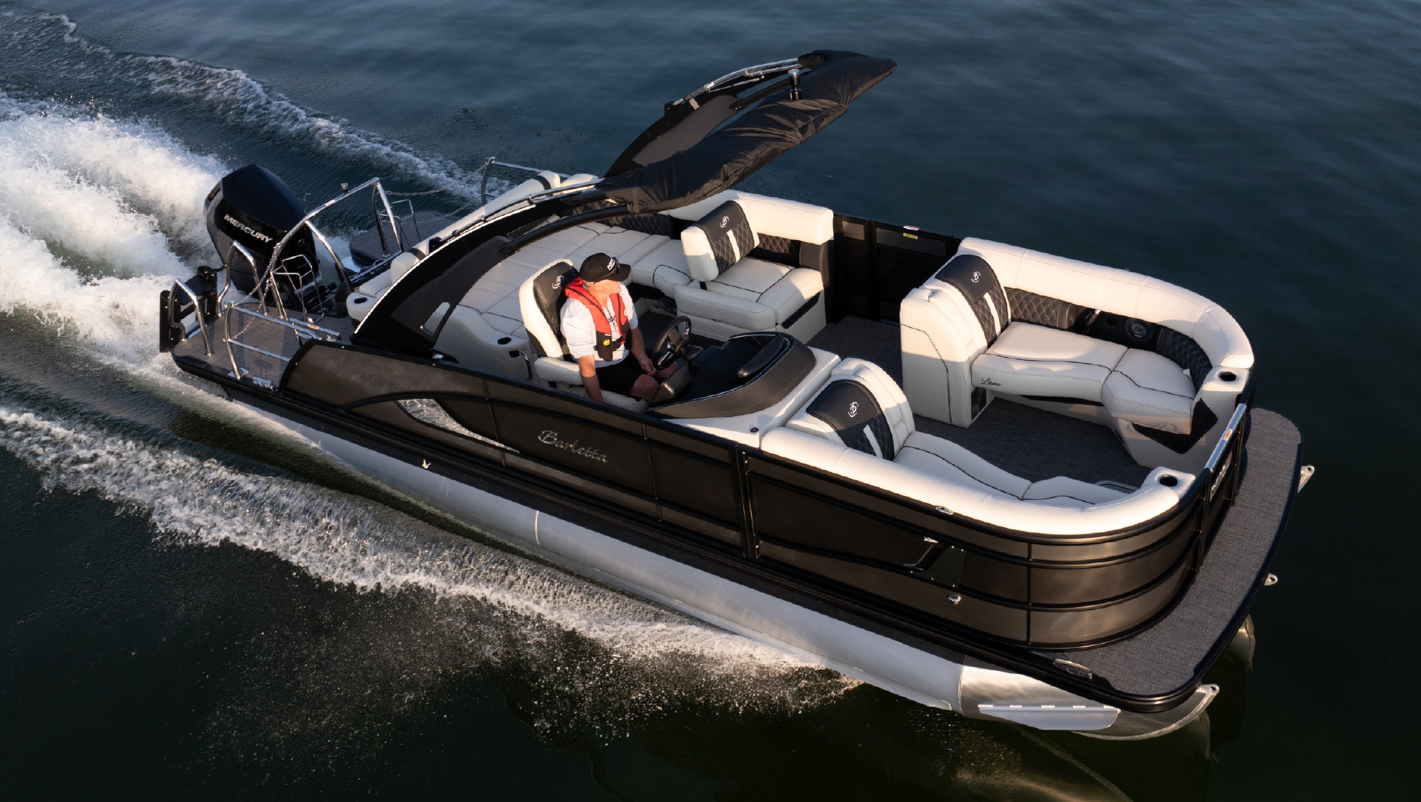 Aerial top high angle view of a man in a driver's seat of a white and black colored Barletta Lusso L23UA pontoon motorboat vehicle as it is cruising forward in motion across a body of water