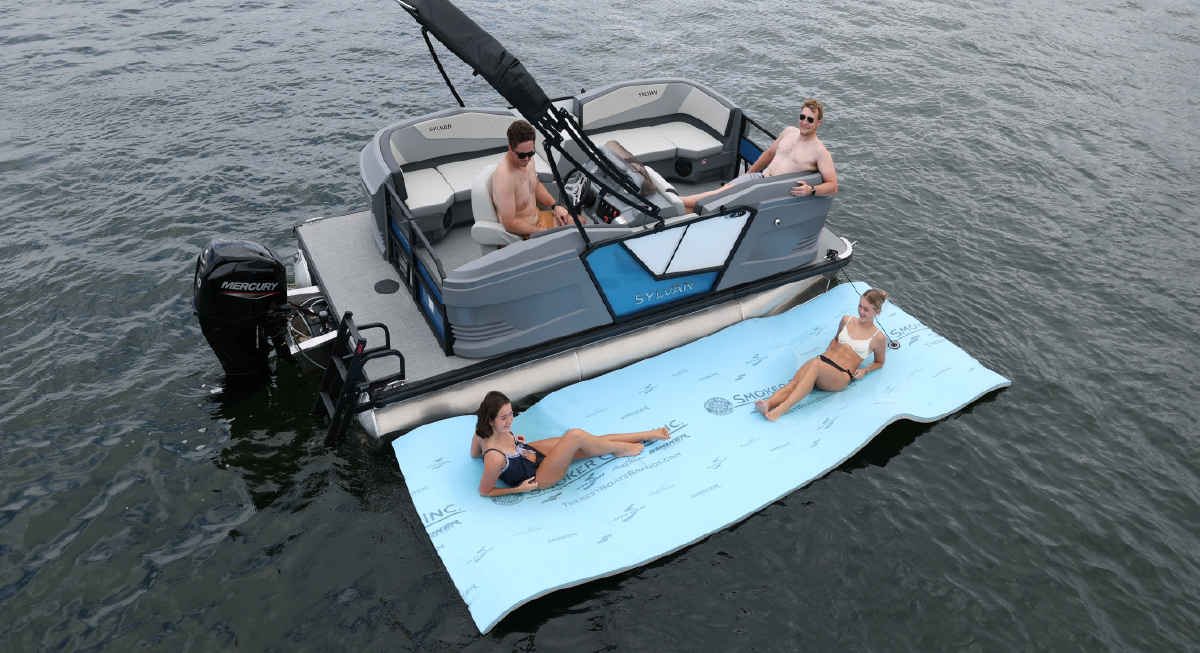 A group of four people as two of the individuals are inside the Sylvan A-16 LZ pontoon motorboat vehicle while the two other individuals are on a Smoker Craft water material floatie sheet of some kind nearby the pontoon motorboat vehicle