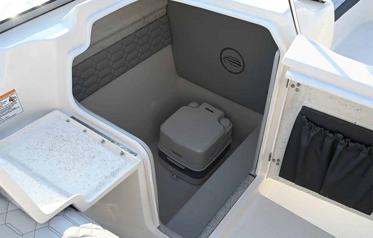A portable marine toilet installed in a dedicated storage compartment with grey honeycomb-patterned upholstery on a modern deck boat.