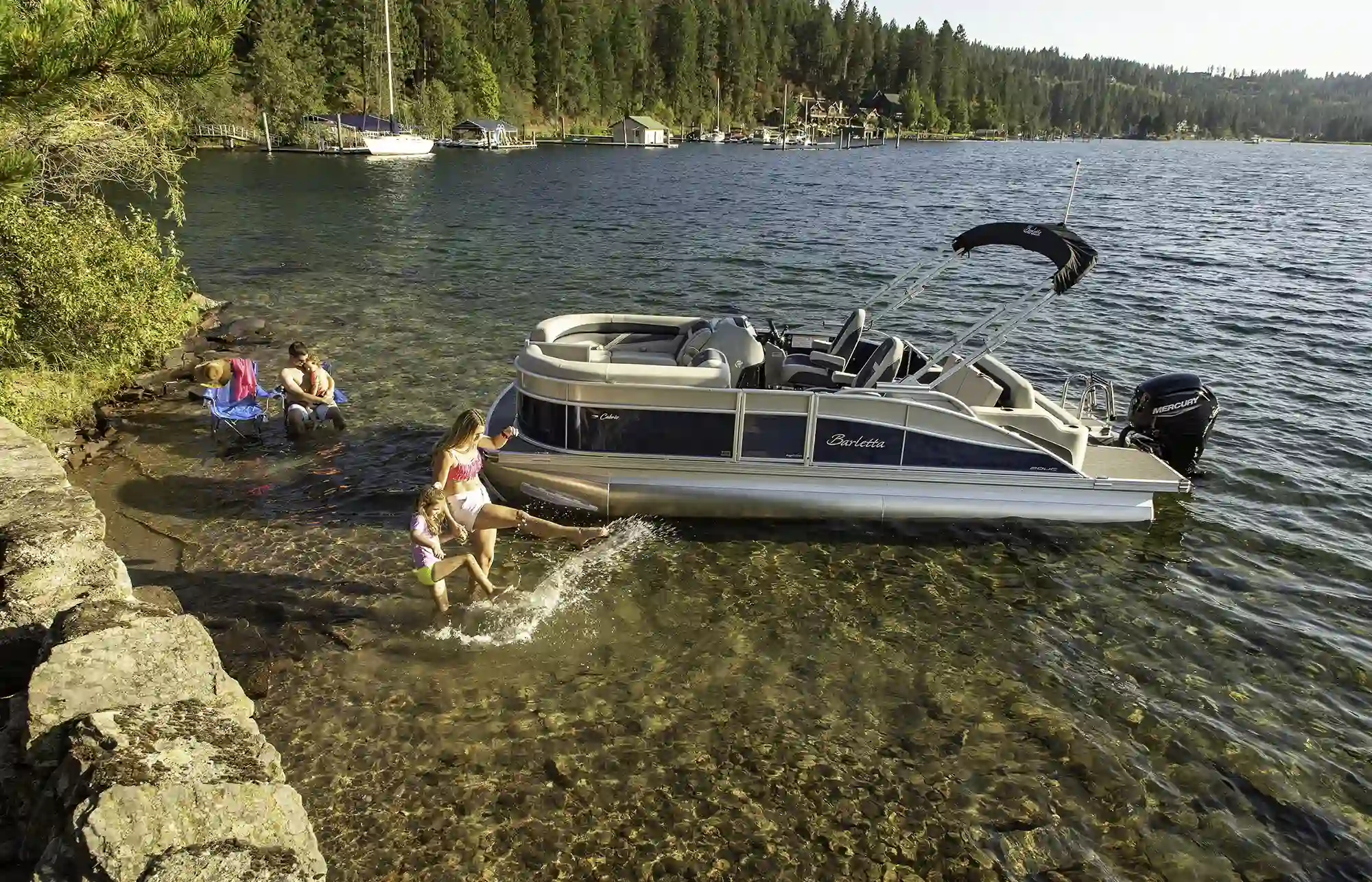 A family playing in the clear shallow water next to their Barletta pontoon boat at a scenic rocky shoreline.