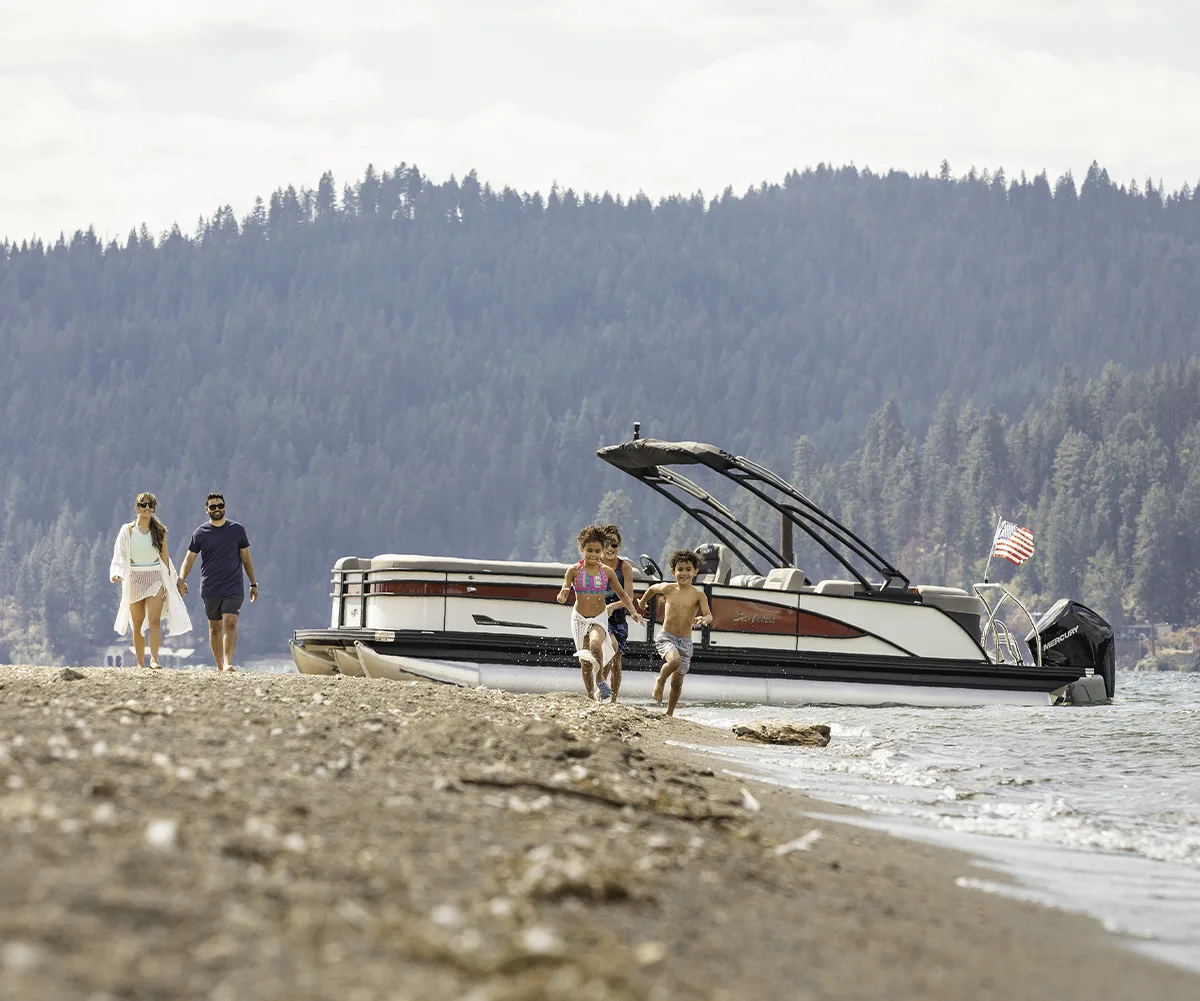 A happy family with young children running along a sandy beach toward the water with their Barletta pontoon boat anchored in the background.