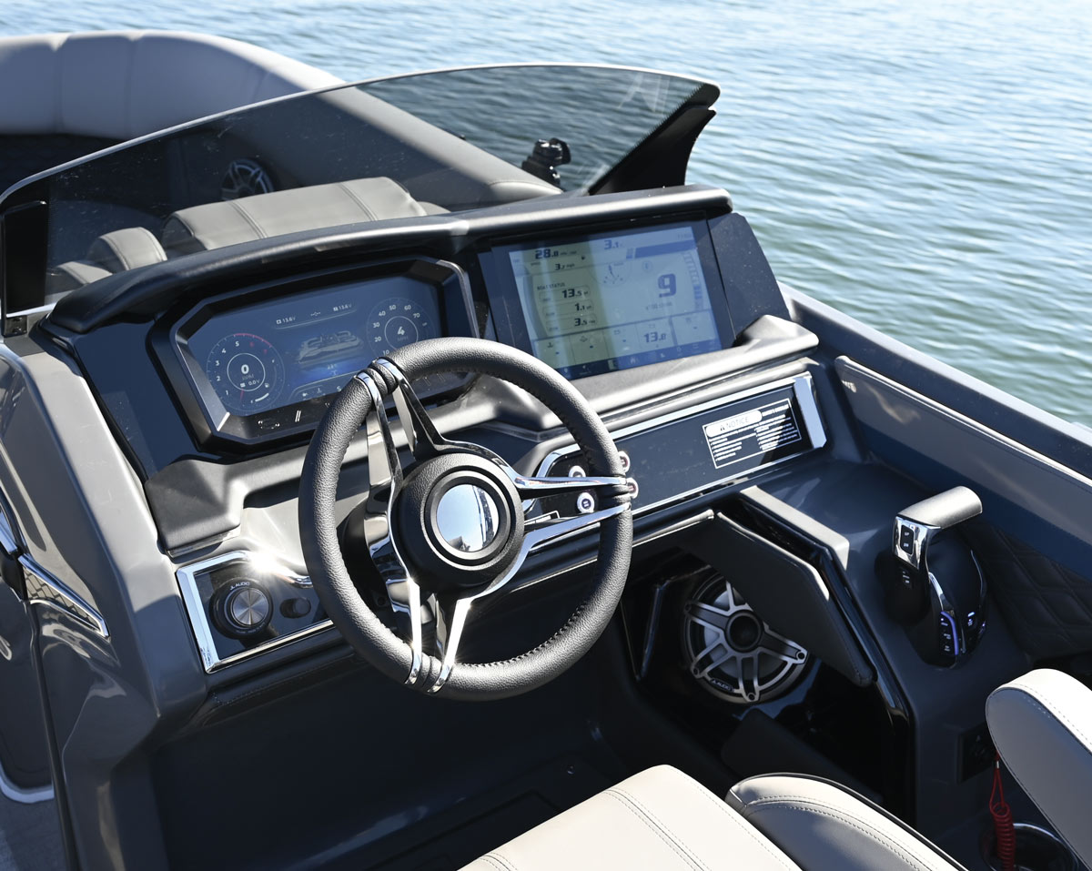 The modern cockpit of a Godfrey pontoon boat featuring a black leather steering wheel, dual digital touchscreen displays, a JL Audio sound system, and a side-mount throttle.