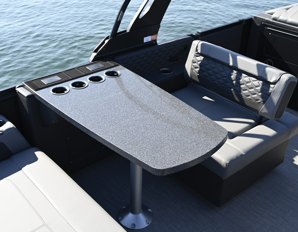 Interior detail of a gray granite-style pedestal table on a boat deck. The table includes four integrated stainless steel cup holders and is flanked by quilted gray seating.