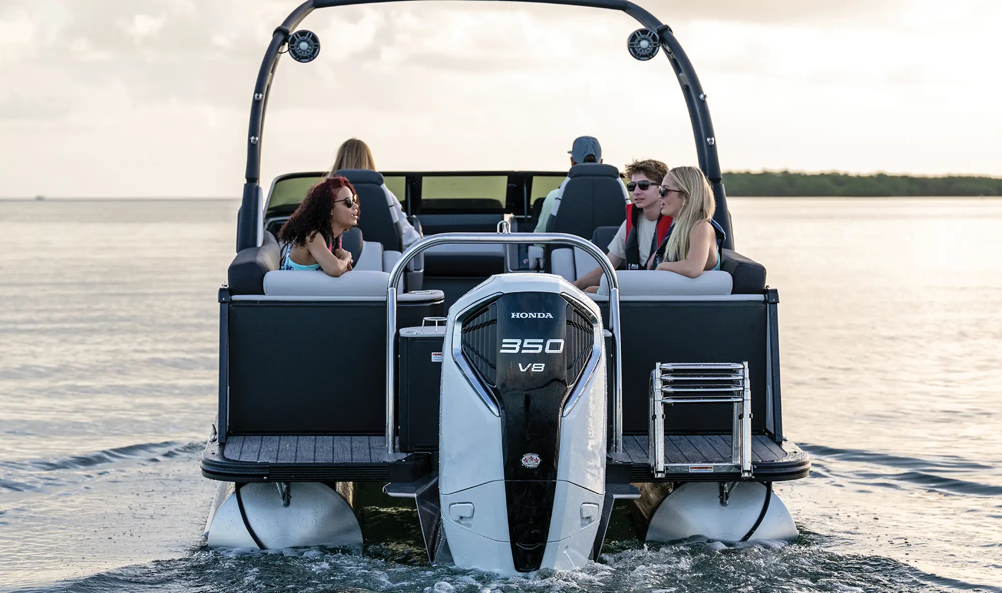 A direct rear view of a modern pontoon boat featuring a powerful white Honda 350 V8 outboard engine. The boat has a black wakeboard tower with integrated speakers and passengers seated in the lounge area.
