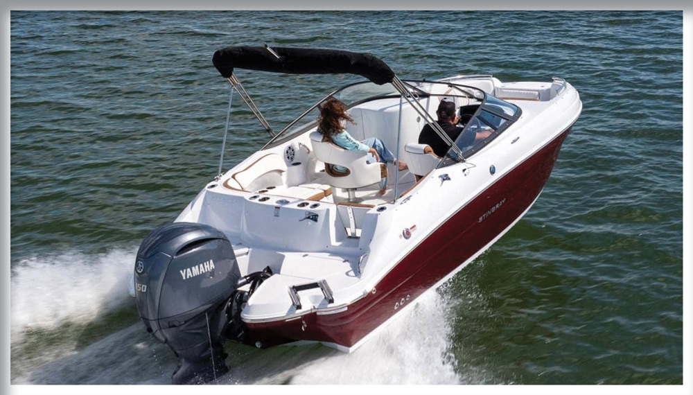 An aerial rear view of a red and white Stingray deck boat cruising at high speed. It is powered by a grey Yamaha outboard engine and features a white interior with a black bimini top.