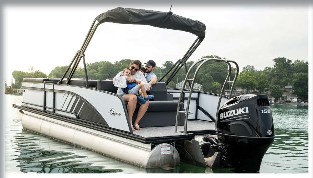 A stationary Qwest pontoon boat on clear water. The boat is equipped with a black Suzuki 150 four-stroke outboard engine. A family is sitting together on the rear bench seat.