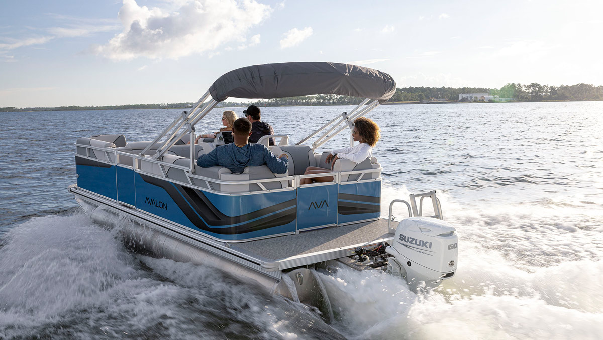 An Avalon pontoon boat with blue and black exterior panels speeding across a lake. A white Suzuki 60 outboard engine is visible at the stern, and four passengers are enjoying the ride under a grey bimini top.