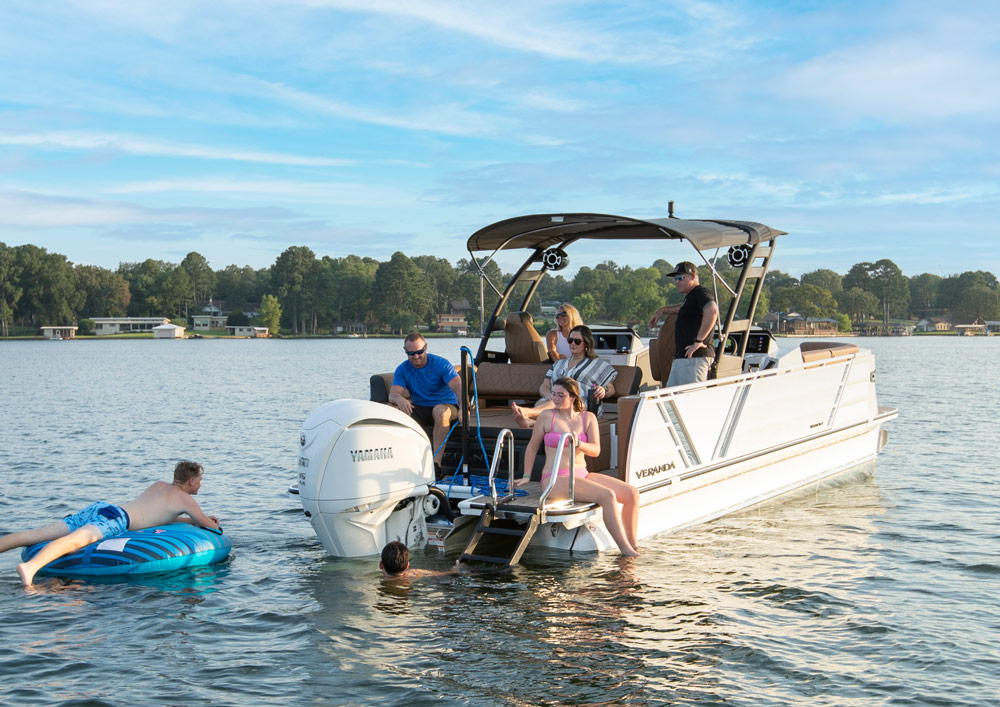 A Veranda pontoon boat anchored on a calm lake. A large white Yamaha outboard engine is mounted on the back. People are relaxing on the deck, and one person is floating nearby on a blue inflatable tube.