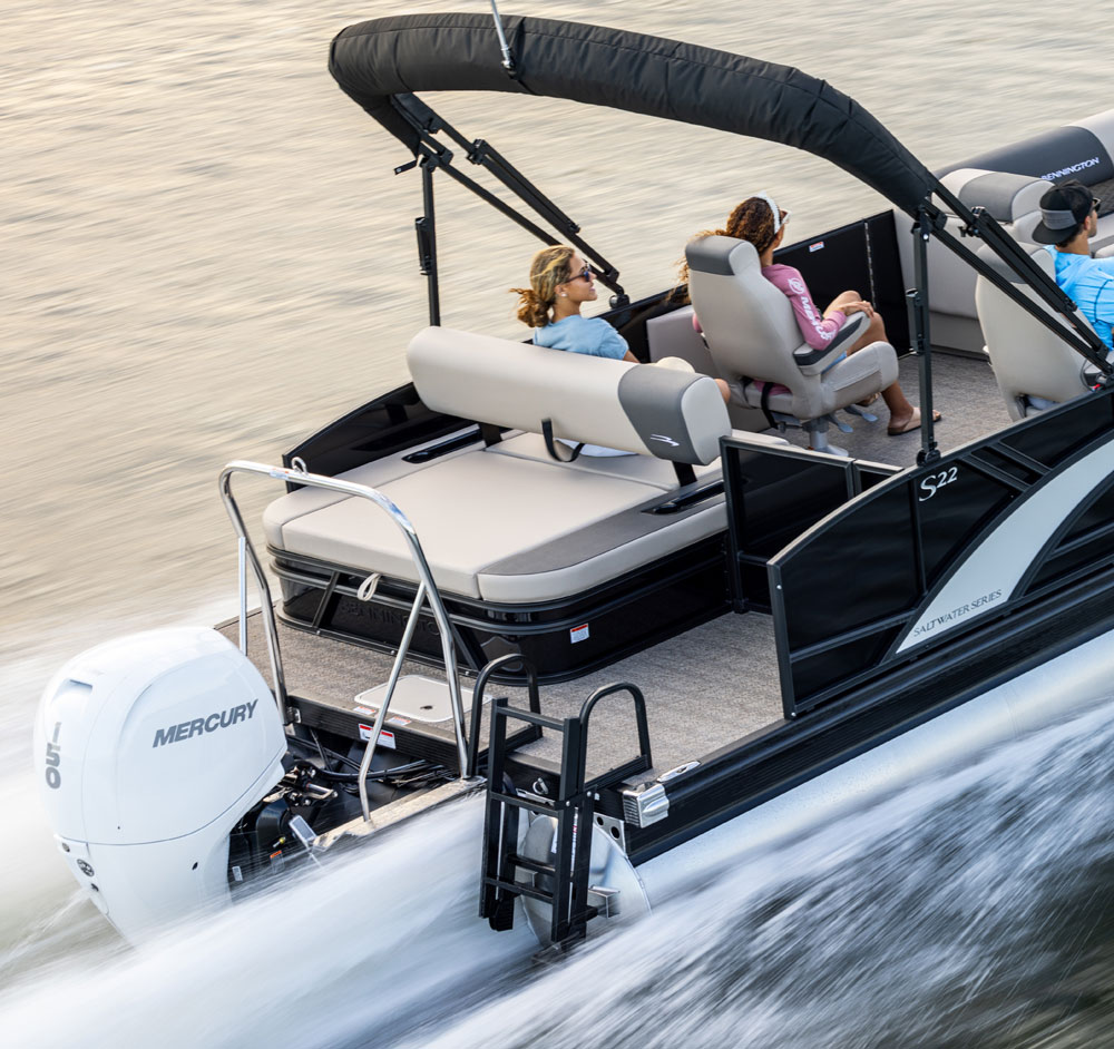 A rear-angle view of a Bennington S22 Saltwater Series pontoon boat cruising on the water. It is powered by a white Mercury 150 outboard engine and features tan and grey upholstery with a black bimini top.
