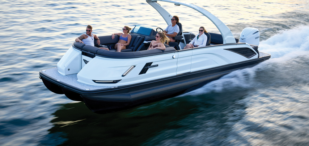 A white Bennington pontoon boat with a large white sport arch travels at high speed. Five passengers enjoy the wrap-around bow seating and comfortable helm chairs.