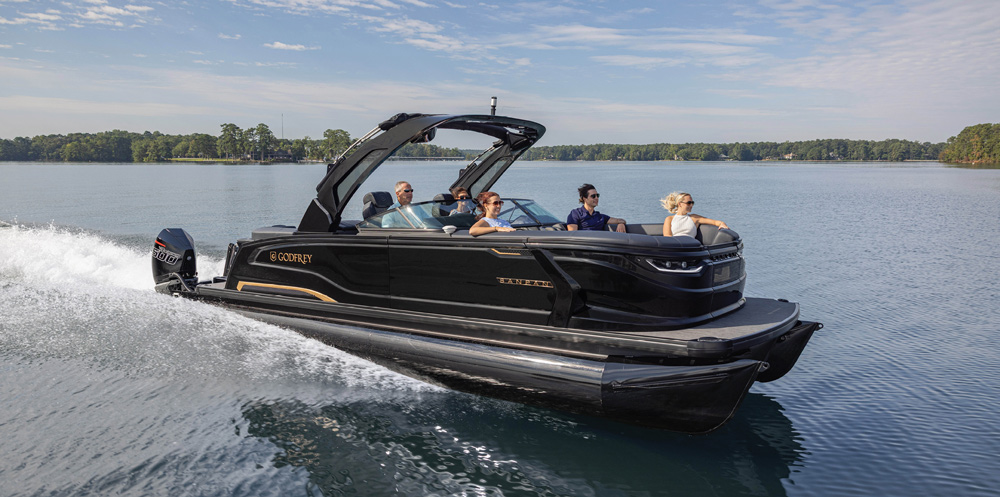 A premium black Godfrey Sanpan pontoon boat with gold accents speeds across the lake. The design features a large black sport tower and integrated windshield for the crew.