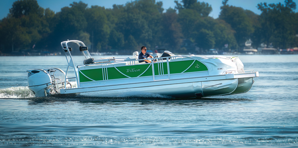 A green and white JC TriToon boat with dual Suzuki outboard engines. The captain drives the vessel, which features high-profile railings and a sturdy white tow arch.