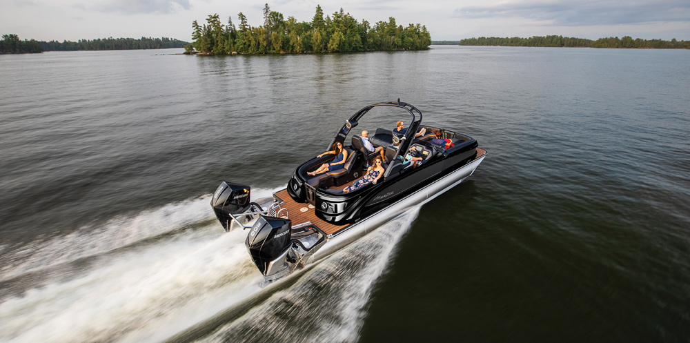 An aerial view of a black Manitou pontoon boat with dual 300hp Mercury engines. The wood-accented deck is filled with passengers relaxing as the boat cruises.