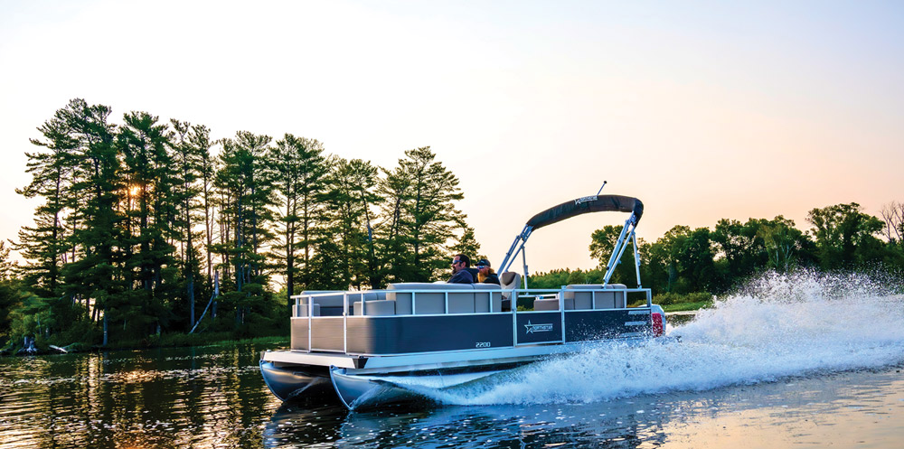 A Northstar 2200 pontoon boat cruises at sunset. The dark grey exterior contrasts with the white wake as two passengers sit near the helm under a black bimini.