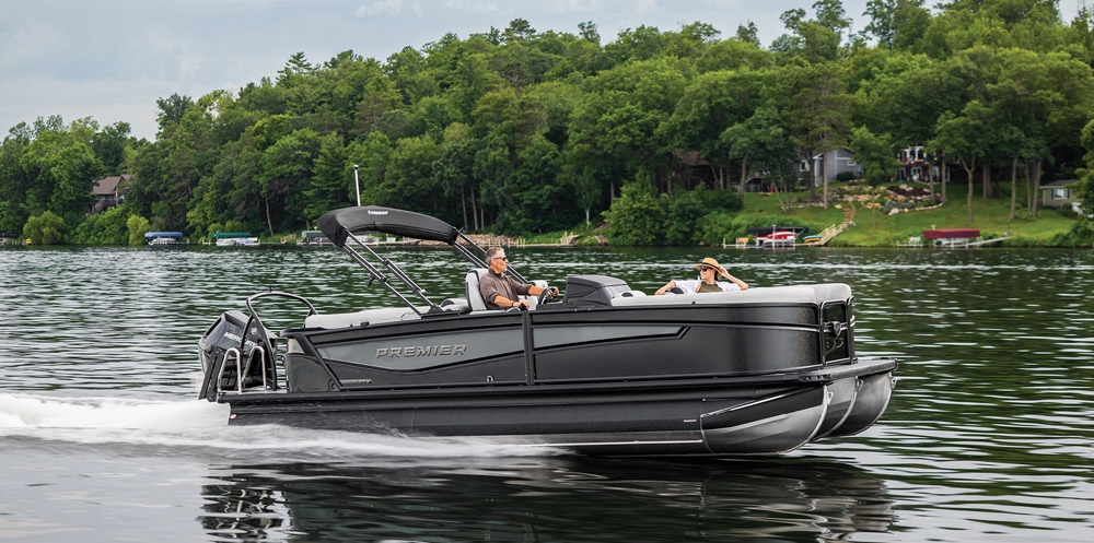 A sleek, all-black Premier pontoon boat glides across a lake. The minimalist design includes a grey interior and a single passenger relaxing in the bow.