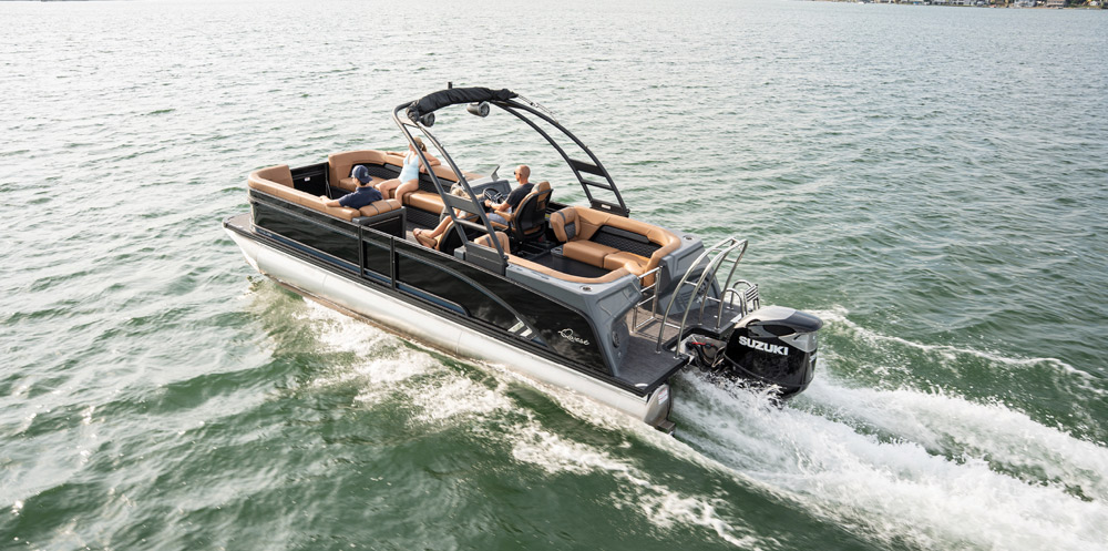 A Qwest pontoon boat with a tan and black interior speeds across the water. It is powered by a black Suzuki outboard and features a high-profile black sport tower.