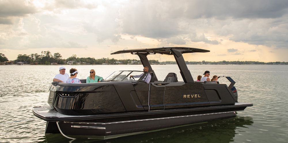 A modern black Revel pontoon boat with an integrated hardtop T-top. Several passengers sit in the spacious lounge areas while the boat is stationary on calm water.
