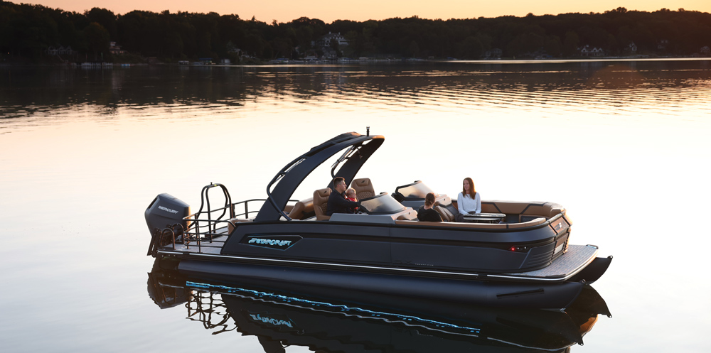 A Starcraft pontoon boat with a grey exterior and tan diamond-stitched seating. A family relaxes on board at sunset under the shade of a curved black tower.