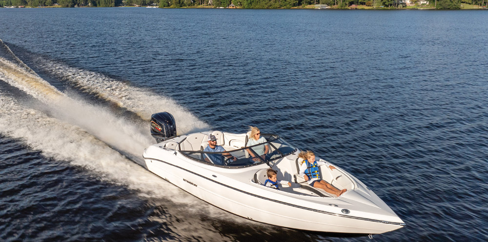 A white Stingray deck boat with a black Mercury engine creates a large wake. Four passengers, including a child, are seated in the bow and cockpit.
