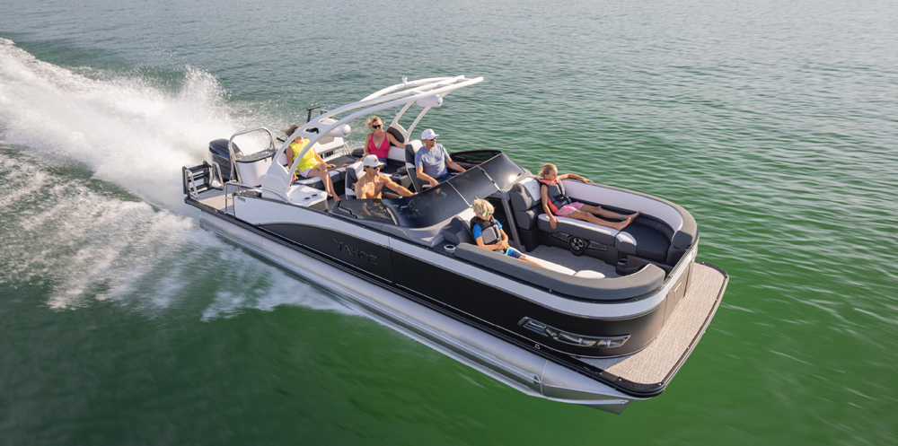 A high-angle view of a Tahoe pontoon boat with a white sport arch. A family is spread out across the black and white upholstered loungers.