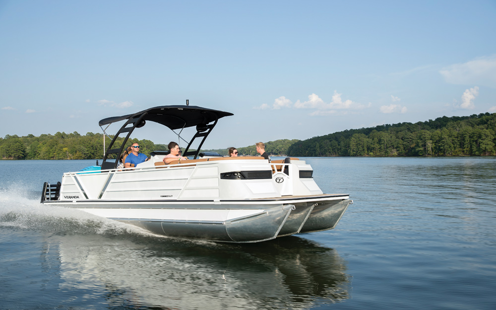 A white Veranda pontoon boat with a black bimini cruises on a calm lake. The boat features a modern, clean exterior and tan interior seating.