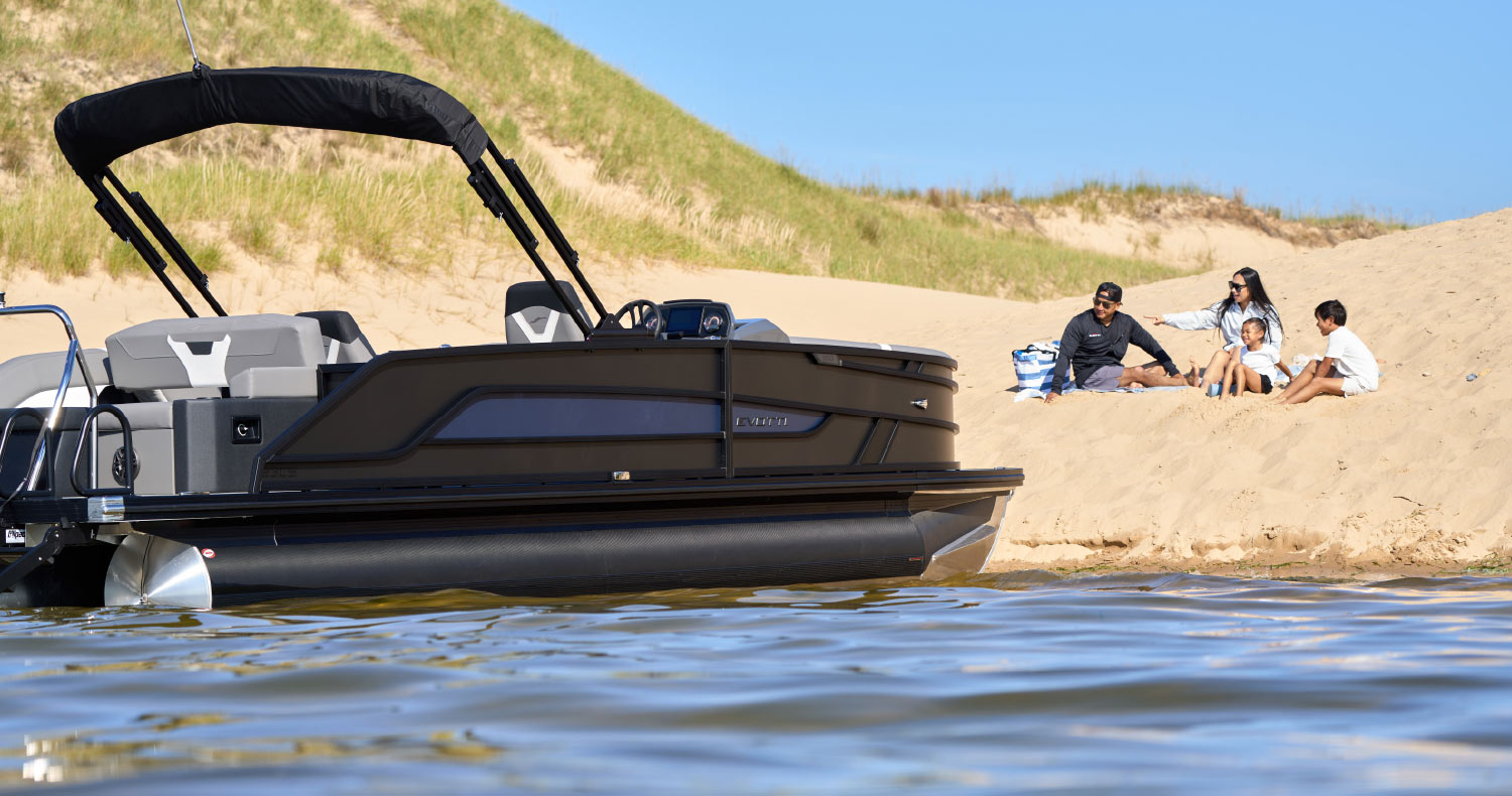 A family of four sits on a sandy dune next to a black pontoon boat anchored in shallow, clear water.
