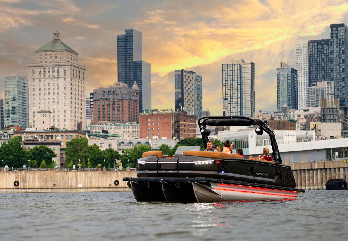 A modern black pontoon boat with glowing red accent lights cruises on a river in front of a large city skyline at dusk.