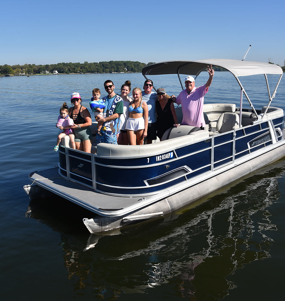 A smiling multi-generational family of nine, including small children, poses together on a blue and white Starpoint pontoon boat on a calm lake under a clear blue sky.