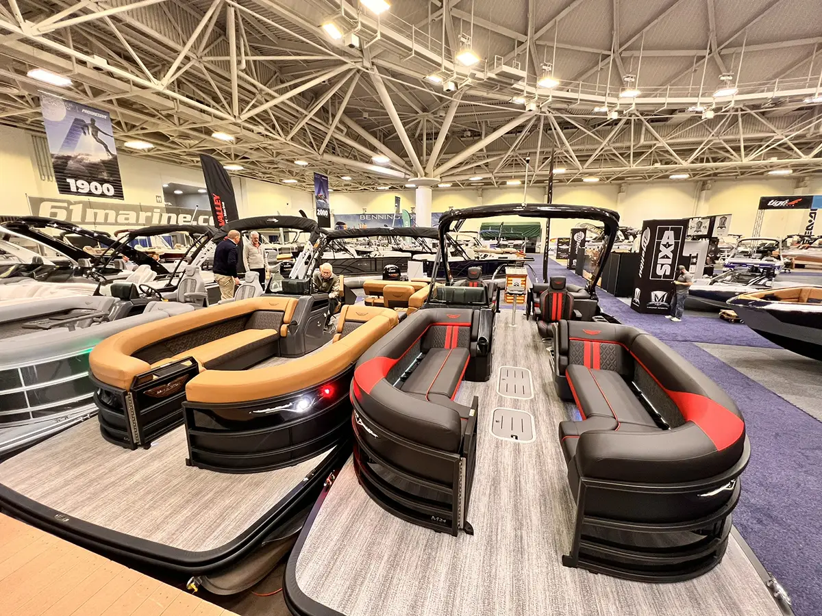 The interior of a large boat showroom featuring several modern pontoon boats with luxury seating, including models with tan and black-and-red upholstery.