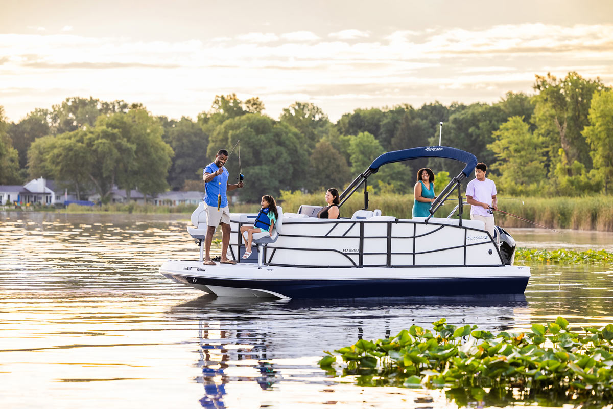 A family of five enjoys fishing from a white Hurricane deck boat on a peaceful lily pad-filled lake during a golden sunset.