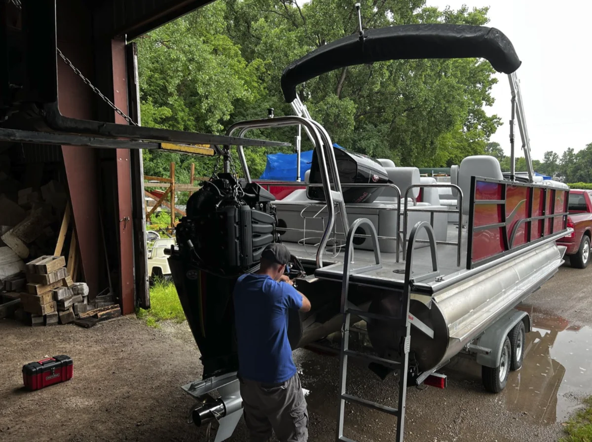 A mechanic in a blue shirt works on a large black outboard engine mounted to a red and silver pontoon boat parked on a trailer outside a service garage.