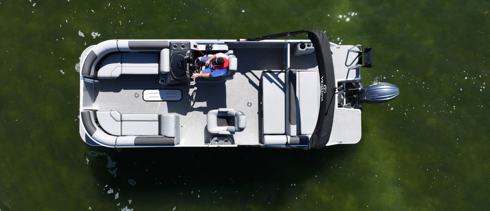 Bird's-eye view of a light grey Veranda pontoon boat on green water showing a central walkway and a driver in a red life vest.