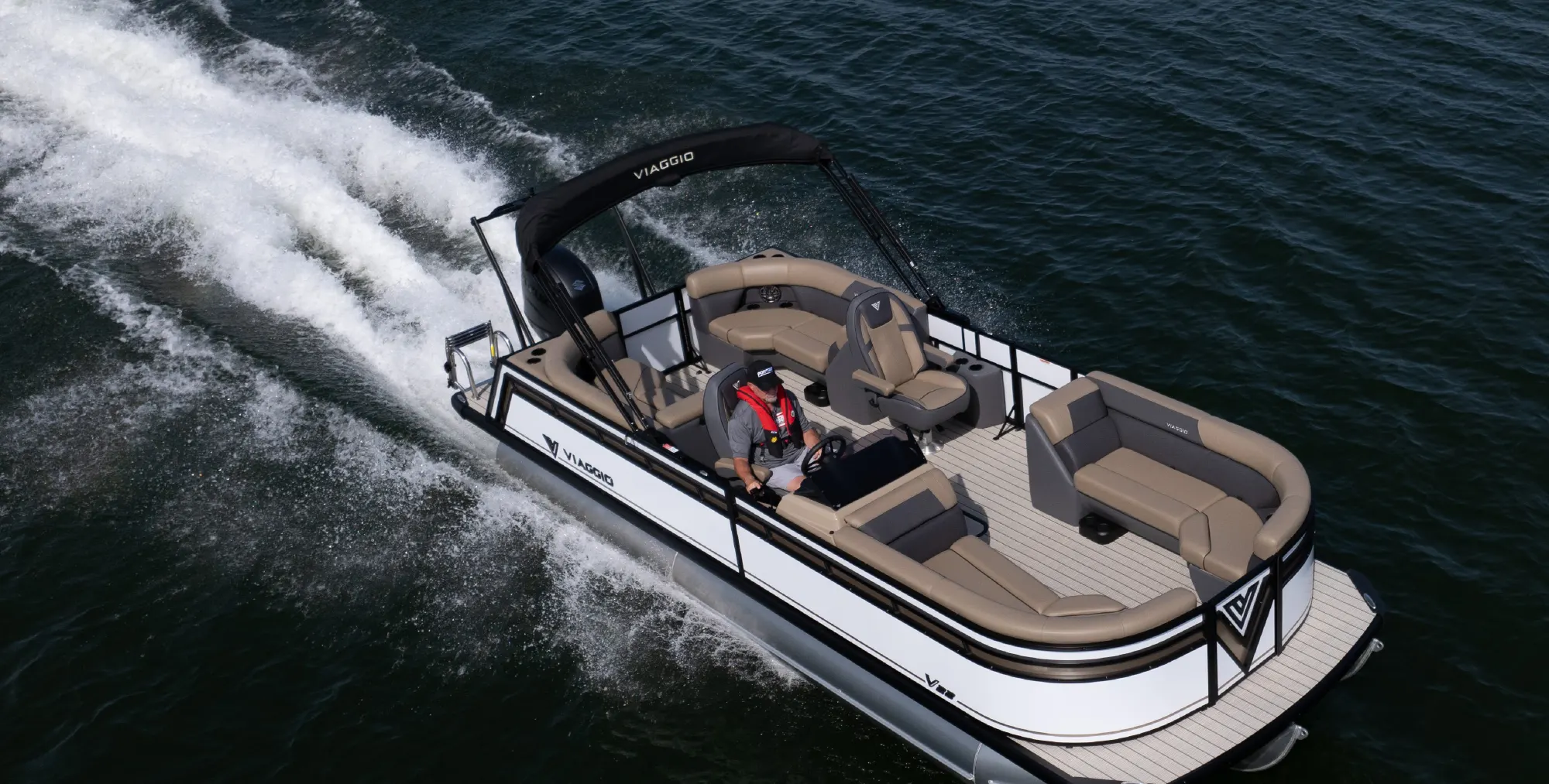 A white and black Viaggio pontoon boat cruising fast on dark water, piloted by a driver in a red life vest.
