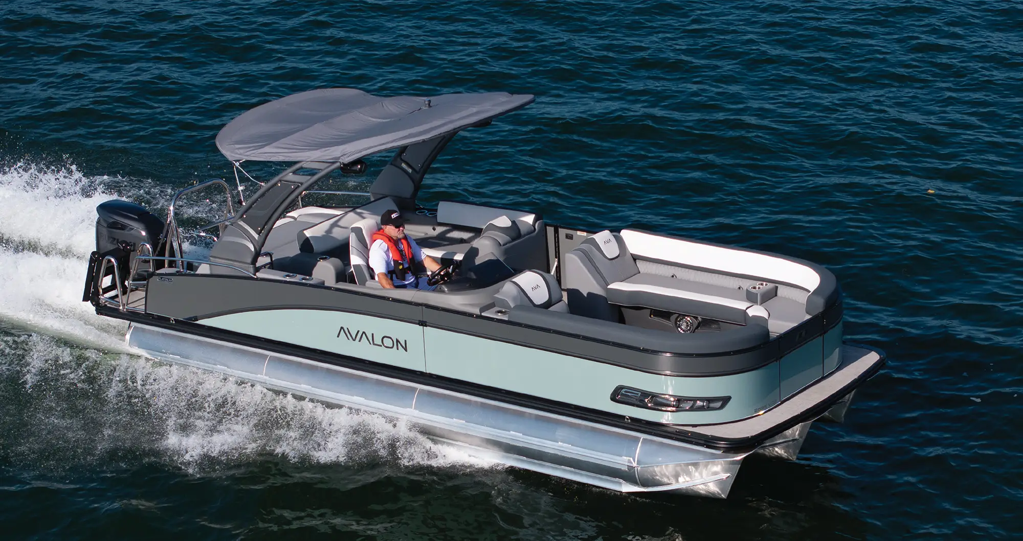 High-angle side view of a sage green and charcoal Avalon pontoon boat cruising on the water. It features a large gray bimini top and a driver wearing a life vest at the helm.