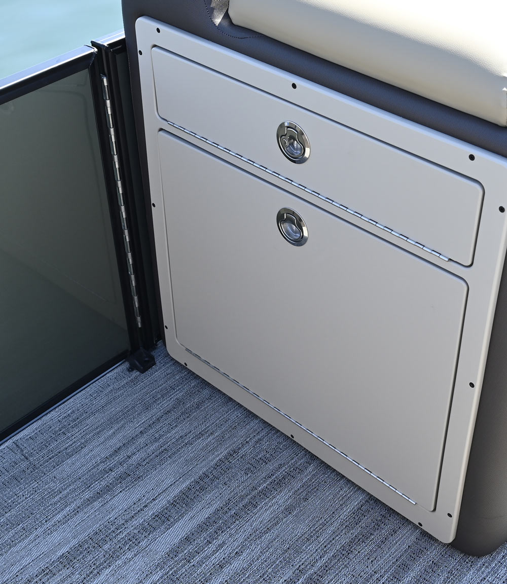 Close-up of a light gray pontoon boat storage console with two closed doors featuring circular chrome latches, set against gray woven vinyl flooring.